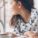 Young girl gazing out window, cup on table reflecting peaceful home ambiance for HVAC comfort tips.