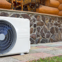 Outdoor air conditioning unit against a stone wall, part of Chad's AC Direct HVAC services in a log cabin setting.