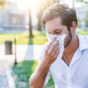 Young man wiping his face with a tissue in a bright outdoor setting, reflecting on HVAC maintenance and summer AC tips.
