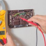 Technician measuring thermostat voltage with multimeter; shows exposed wiring for HVAC maintenance at Chad's AC Direct.