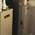 Heating units in a utility room; includes beige equipment, piping, and brand labels by Chad's AC Direct.