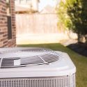 Air conditioning unit in a residential setting with grass and brick wall under sunny skies. Chad's HVAC services.